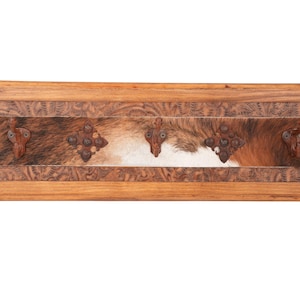 May include: Wooden wall-mounted coat rack with a rectangular design. The frame is made of light brown wood with carved floral patterns. The center features a cowhide panel with brown, white, and tan fur. Includes three decorative metal hooks.