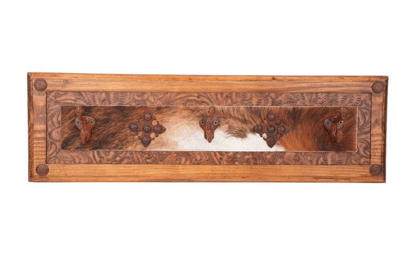 May include: Wooden wall-mounted coat rack with a rectangular design. The frame is made of light brown wood with carved floral patterns. The center features a cowhide panel with brown, white, and tan fur. Includes three decorative metal hooks.
