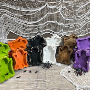 May include: A collection of colorful, ghost-shaped trays in various colors, including green, orange, black, white, brown, and purple. The trays are arranged on a light-colored surface with decorative spiders and a white spiderweb backdrop, perfect for Halloween.