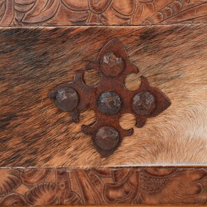 May include: Close-up of a rustic, textured surface featuring a cowhide pattern in shades of brown and white. A decorative, rusty metal accent in a cross shape is riveted to the hide. The top and bottom edges have carved leather details.