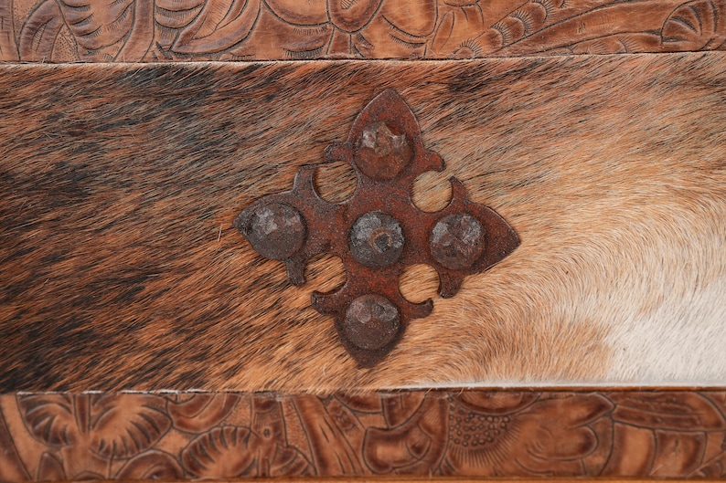 May include: Close-up of a rustic, textured surface featuring a cowhide pattern in shades of brown and white. A decorative, rusty metal accent in a cross shape is riveted to the hide. The top and bottom edges have carved leather details.