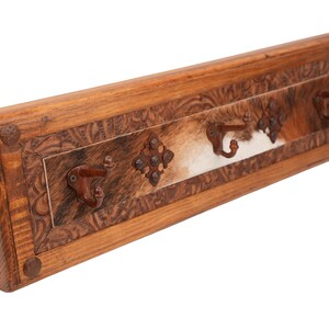 May include: A rustic wooden coat rack with three metal hooks. The rack features a carved border and a central panel with a cowhide design. Decorative metal accents add to the western aesthetic.