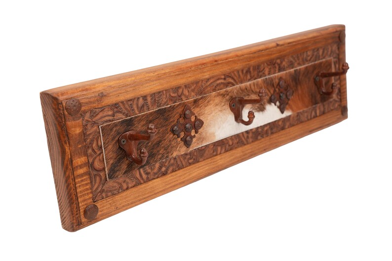 May include: A rustic wooden coat rack with three metal hooks. The rack features a carved border and a central panel with a cowhide design. Decorative metal accents add to the western aesthetic.