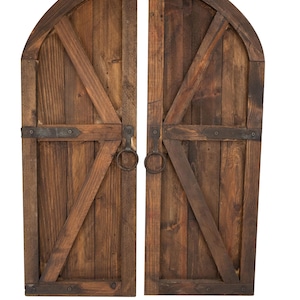May include: Two rustic wooden arched doors with a dark brown finish. Each door features diagonal wooden supports, metal hinges, and a round metal handle. The doors are designed to be decorative.