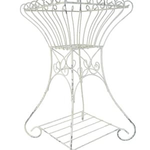 May include: Off-white, wrought iron plant stand with a decorative, openwork design. The stand features a square base with a slatted shelf and curved legs. The top has a basket-like shape with decorative swirls.