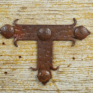 May include: A rusty metal T-shaped hinge with decorative scrolls and rounded edges. The hinge is attached to a weathered wooden surface.