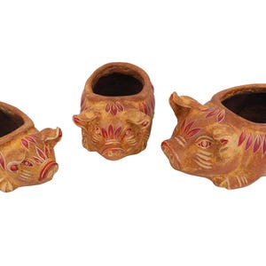 May include: Three pig-shaped planters made of brown clay with hand-painted red and white leaf-like patterns on the ears, faces, and backs. Each planter features a hollow center for holding small plants, with distinct snout and ear details.