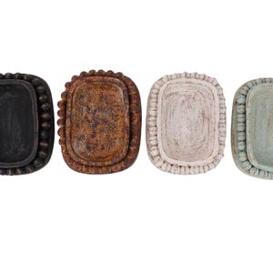 May include: Four rectangular ceramic trays arranged in a row, each featuring a raised beaded border. The trays are finished in distinct colors: matte black, textured brown, off-white with a weathered appearance, and a pale seafoam green.
