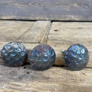 May include: Three hammered metal knobs with a rustic blue-green finish. The knobs have a round shape and a textured surface.