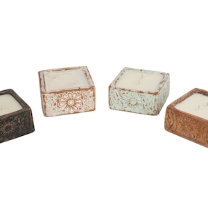 May include: Four square candles in decorative holders. The holders are in shades of brown, white, and blue, with floral patterns. Each candle has a white wax and two wicks.