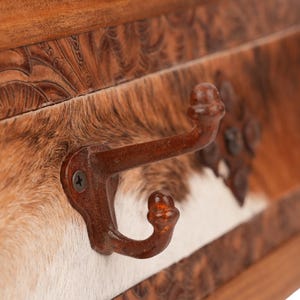 May include: A rustic wall-mounted coat rack with three metal hooks. The rack features a cowhide panel with brown, tan, and white markings, and a carved wooden frame. The hooks are a dark brown color.
