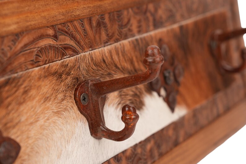 May include: A rustic wall-mounted coat rack with three metal hooks. The rack features a cowhide panel with brown, tan, and white markings, and a carved wooden frame. The hooks are a dark brown color.