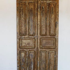 May include: A pair of antique wooden doors with a distressed finish. The doors are made of wood panels with a raised design. The doors are painted in a light brown color with a white wash.