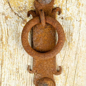 May include: A rusty metal door knocker with a circular ring handle and ornate details. The knocker is attached to a wooden door with white paint.
