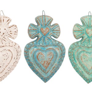 May include: Three heart-shaped wall hangings in off-white, turquoise, and teal. Each has a heart design in the center and a cross-like shape at the top. The hangings have a distressed, rustic appearance.