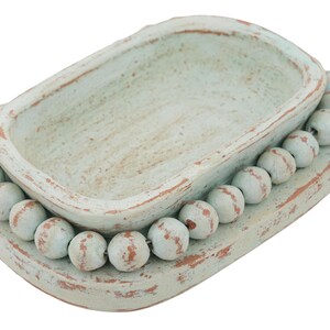 May include: A distressed, rectangular, light blue decorative tray with a central basin. The tray is encircled by a row of white, round beads with brown accents, creating a rustic, farmhouse aesthetic. The tray is made of wood.