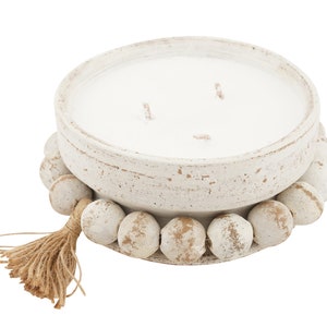 May include: White ceramic candle holder with a white candle and a wooden bead trim. The holder has a natural jute tassel.