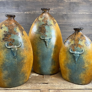 May include: Three oval-shaped ceramic vases with a mottled finish in shades of mustard yellow, deep blue, and rust brown. Each vase features a centered, raised relief sculpture of a bull skull on the front and a narrow neck opening.