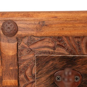 May include: Close-up of a wooden furniture corner. The frame is made of light brown wood with a dark brown, textured metal rivet. The center features a brown leather panel with carved floral designs and a section of brown and tan animal hide.