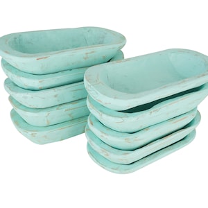 May include: Stacks of small, aqua-colored wooden bowls with a distressed finish. The bowls are oval-shaped and appear to be handmade, with visible wood grain and a slightly worn appearance. The bowls are stacked in two groups.