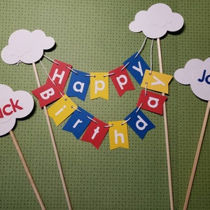 May include: A colorful birthday cake topper set with cloud-shaped decorations and a banner that spells "Happy Birthday." The clouds have the names "Zack" and "Joey" on them. The banner is red, yellow, and blue.