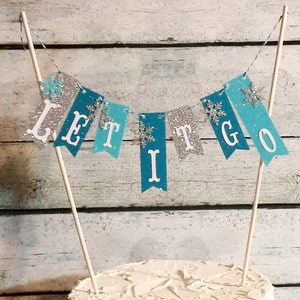 May include: A cake topper with a banner that reads "LET IT GO". The banner has silver and teal pennants with white letters and snowflake accents. The banner is strung between two wooden dowels.
