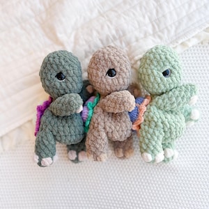 Puede incluir: Tres peluches de tortuga hechos a mano con ganchillo en tonos verdes y marrones. Cada tortuga tiene ojos de seguridad negros y un caparazón colorido. Los peluches se exhiben sobre una superficie blanca texturizada.