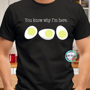 Puede incluir: Camiseta negra con la frase "You know why I'm here" y tres ilustraciones de dibujos animados de huevos duros. Los huevos tienen claras de huevo blancas y yemas amarillas. Un pequeño logotipo está en el lado derecho.