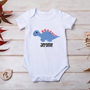 May include: White baby onesie featuring a blue dinosaur and the name "Jerome" printed on the front.