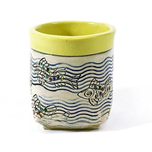 May include: A yellow ceramic mug with a white and blue design of fish swimming in waves. The fish are decorated with colorful dots.