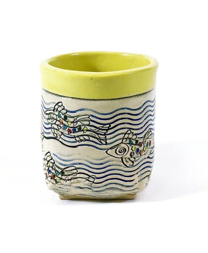 May include: A yellow ceramic mug with a white and blue design of fish swimming in waves. The fish are decorated with colorful dots.