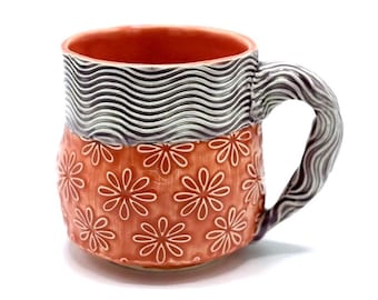 May include: A handmade ceramic mug with a wavy pattern in black and white on the top and a floral pattern in white on a red background. The mug has a handle with a wavy pattern.
