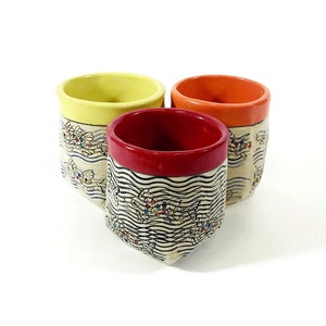May include: Three ceramic mugs with a white background and black and white wave pattern. The mugs have different colored rims: yellow, red, and orange.