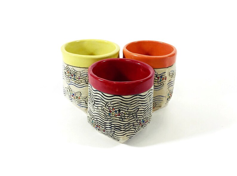 May include: Three ceramic mugs with a white background and black and white wave pattern. The mugs have different colored rims: yellow, red, and orange.