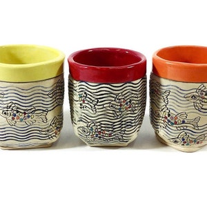May include: Three ceramic mugs with a white background and blue wavy lines. Each mug has a different colored rim: yellow, red, and orange. The mugs are decorated with colorful fish swimming in the wavy lines.