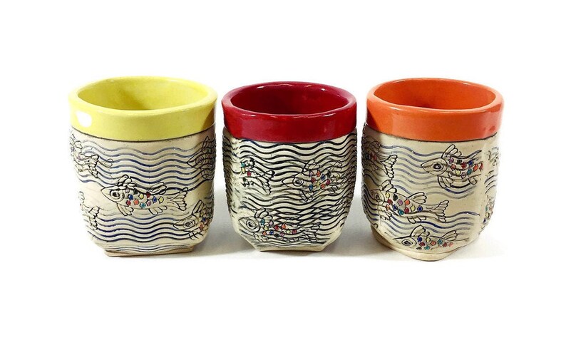 May include: Three ceramic mugs with a white background and blue wavy lines. Each mug has a different colored rim: yellow, red, and orange. The mugs are decorated with colorful fish swimming in the wavy lines.