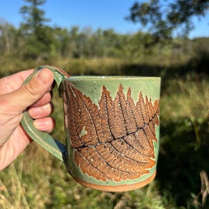 May include: Handmade ceramic mug with a green glaze and a detailed brown fern design. The mug has a large handle and is held against a backdrop of green grass and a blue sky. A unique piece for enjoying your favorite beverage.