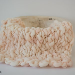 May include: A hand-knitted, light peach-colored basket with a chunky, textured design. The basket's sides are made of thick, looped yarn, creating a soft, tactile appearance. The interior is a lighter shade, providing contrast.