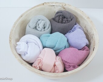 May include: A rustic, light-colored wooden bowl filled with rolled fabric in various pastel colors. The fabrics include white, light gray, teal, lavender, pink, and mauve. The bowl is set against a white background.