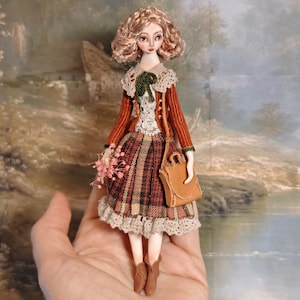 May include: A small doll with curly light brown hair, wearing a rust-colored ribbed cardigan over a white lace-trimmed top and a plaid skirt with a lace hem. The doll carries a small tan bag and holds a tiny bunch of pink flowers, wearing brown boots.