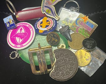 May include: A collection of assorted metal pins, keychains, and tokens including a butterfly pin, a round pink Missionary Union pin, a blue World War II pin, a gold belt buckle, a shark tooth, and a brown Oreo cookie-shaped pin.