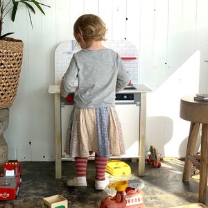May include: A young child playing with a toy kitchen set. The kitchen is white with a black oven and a red knob. The child is wearing a grey long-sleeved shirt and a multi-colored skirt. The child is standing in front of the kitchen and is looking at the oven. There are other toys on the floor, including a red fire truck and a yellow toy.