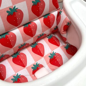 May include: A white high chair with a pink and white checkered fabric seat cushion featuring a strawberry pattern. The cushion has a built-in headrest and a removable tray.
