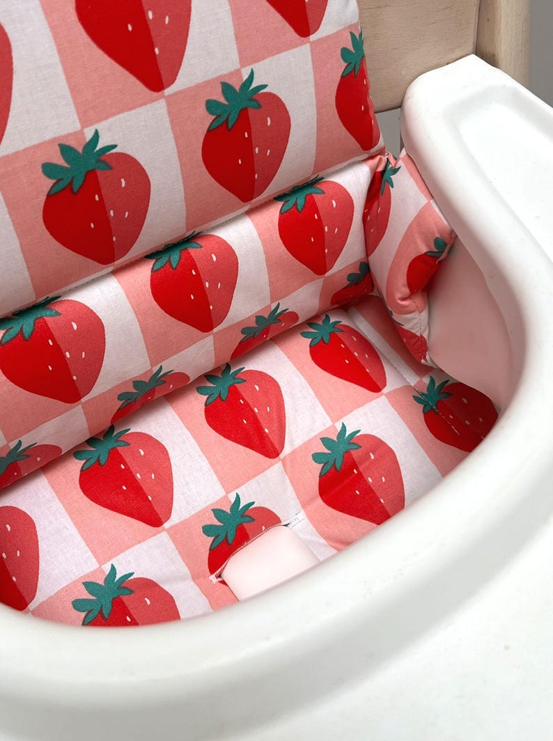 May include: A white high chair with a pink and white checkered fabric seat cushion featuring a strawberry pattern. The cushion has a built-in headrest and a removable tray.