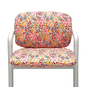 Könnte beinhalten: Weißer Metallstuhl mit floralem Muster auf Sitzfläche und Rückenlehne. Das Muster zeigt rosa, gelbe und rote Blumen mit grünen Blättern auf hellrosa Hintergrund. Der Stuhl hat ein schlichtes, modernes Design.