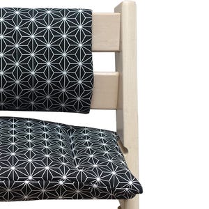 K&ouml;nnte beinhalten: Ein h&ouml;lzerner Hochstuhl mit einem schwarz-wei&szlig;en geometrischen Kissen und einer R&uuml;ckenlehne. Das Muster ist ein sich wiederholendes Design aus wei&szlig;en sternartigen Formen auf schwarzem Hintergrund. Der Stuhlrahmen ist hell und hat eine nat&uuml;rliche Holzfarbe.