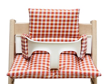 May include: A white high chair with a red and orange checkered cushion and back support. The cushion has a white strap across the back.