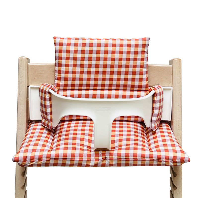 May include: A white high chair with a red and orange checkered cushion and back support. The cushion has a white strap across the back.