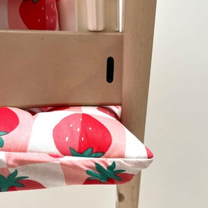 May include: A white high chair with a pink and white cushion featuring a strawberry pattern. The cushion is made of fabric and has a white background with pink and red strawberries.