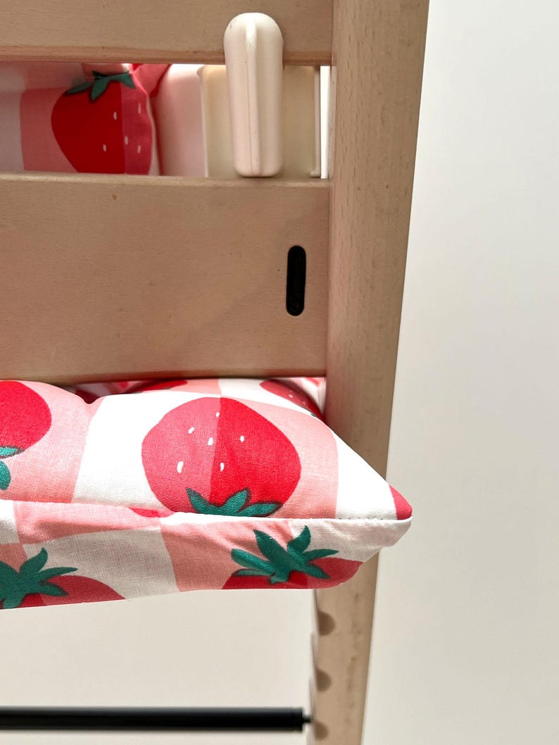 May include: A white high chair with a pink and white cushion featuring a strawberry pattern. The cushion is made of fabric and has a white background with pink and red strawberries.